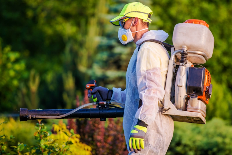 Pest control River Hills in protective gear using a backpack sprayer in a garden.